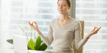 Relaxed woman meditating at workplace, practicing eastern spiritual practices for stress relief and mental health while sitting at desk in front of laptop. Short break in work for strength recovery
