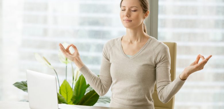 Relaxed woman meditating at workplace, practicing eastern spiritual practices for stress relief and mental health while sitting at desk in front of laptop. Short break in work for strength recovery