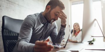 Young handsome frustrated and stressed businessman sitting at the office front a computer and holding head.