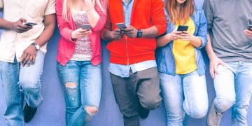 Teenagers texting mobile phone messages leaning on urban wall - Group of multiracial friends using cellular standing outdoors - Concept of students addiction to social network and telephone technology