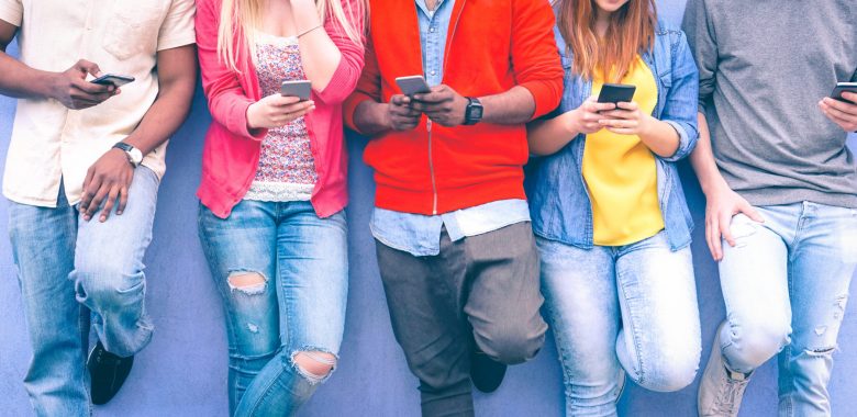 Teenagers texting mobile phone messages leaning on urban wall - Group of multiracial friends using cellular standing outdoors - Concept of students addiction to social network and telephone technology